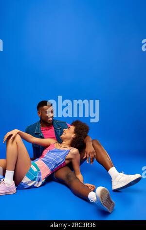 Positive junge afroamerikanische Frau mit kühnem Make-up, die ein Sundkleid trägt, während sie mit ihrem besten Freund im Sommerkostüm posiert und auf blauem Hintergrund sitzt, Stockfoto