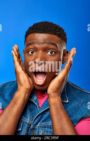 Porträt eines erstaunten jungen afroamerikanischen Mannes mit moderner Frisur, Jeansweste und pinkfarbenem T-Shirt, während er auf die Kamera blickt, isoliert auf blau, Stockfoto