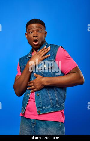 Schockierter junger afroamerikanischer Mann mit moderner Frisur. Er trug eine Jeansweste und ein helles T-Shirt, während er in die Kamera schaute und isoliert auf blau stand. Stockfoto