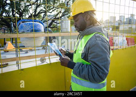Junger Bauingenieur mit gelbem Sicherheitshelm und langer Jacke, stand bei Sonnenuntergang auf der Baustelle mit einem Ordner in der Hand, der das liest Stockfoto