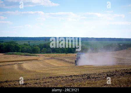 Ein moderner blauer Traktor mit einer gezogenen Scheibenegge und einer Walze pflügt ein Feld, auf dem das Frühjahrsgetreide gerade geerntet wurde. Mittsommer Stockfoto