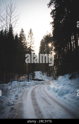 Das Eismärchen in den Beskydy-Bergen. Eine Straße, gesäumt mit Schneehaufen an den Kanten und die Sonne, die bei Sonnenaufgang hinter den Bäumen auftaucht. Stockfoto