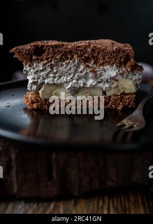 Tiramisu-Kuchen mit Schlagsahne und Banane auf einem schwarzen Teller Stockfoto