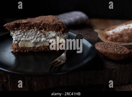 Tiramisu-Kuchen mit Schlagsahne und Banane auf einem schwarzen Teller Stockfoto