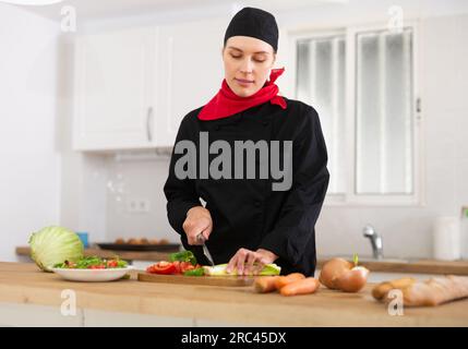 Positive weibliche Küchenchefin in schwarzer Uniform, die Gemüsegericht zubereitet Stockfoto