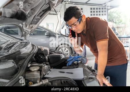 Ein asiatischer Automechaniker, der sein Handy benutzt, während er in seiner Werkstatt an der Reparatur eines Motors arbeitet Stockfoto