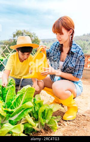 Liebevolle Mutter mit niedlichem Jungen in lässiger Kleidung und Hut auf sandigem Boden, während er bei Tageslicht im Garten üppigen grünen Salat bewässert Stockfoto