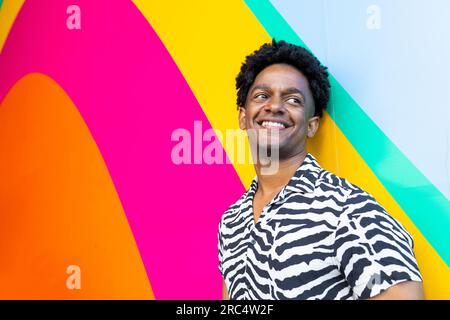 Porträt eines positiven jungen afroamerikanischen Mannes im gestreiften Hemd, der lächelt und wegschaut, während er vor einem bunten Wandhintergrund steht Stockfoto