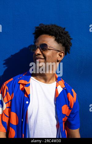 Porträt eines glücklichen afroamerikanischen Mannes in trendiger Sonnenbrille mit lockigem Haar in Sonnenbrille, die an der blauen Wand steht Stockfoto