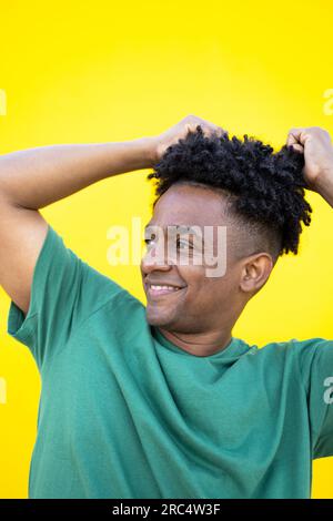 Fröhlicher junger afroamerikanischer Mann in lockigem Haar und lässiger Kleidung mit erhobenen Händen, die das Haar berühren, während er auf gelbem Hintergrund steht und aussieht Stockfoto