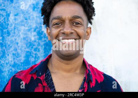 Schnittbild eines fröhlichen afroamerikanischen Mannes mit lockigem Haar, der in die Kamera schaut, während er lächelt und neben der Wand steht, mit blauem und weißem Hintergrund Stockfoto