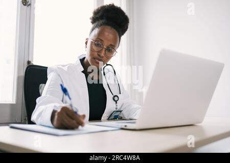 Konzentrierte afroamerikanische Frau in medizinischer Uniform, sitzt am Tisch mit Laptop und schreibt auf Papier Stockfoto