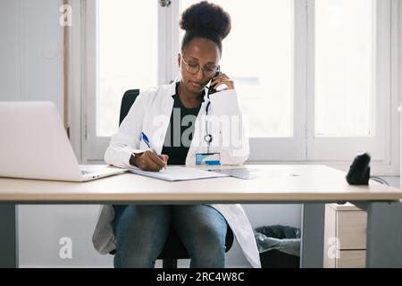 Junge afroamerikanische Ärztin in weißer Uniform, die am Tisch sitzt und telefoniert, während sie in der modernen Klinik Notizen schreibt Stockfoto