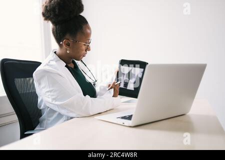 Seitenansicht einer afroamerikanischen Ärztin in Uniform, Brille und Stethoskop, die auf den Bildschirm eines Laptops schaut, während sie am Tisch sitzt und x ra zeigt Stockfoto