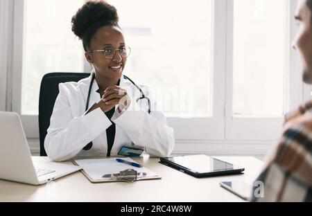 Positive afroamerikanische Ärztin interagiert mit einem nicht erkennbaren männlichen Patienten und schaut sich an, während sie am Tisch mit einem Laptop sitzt Stockfoto