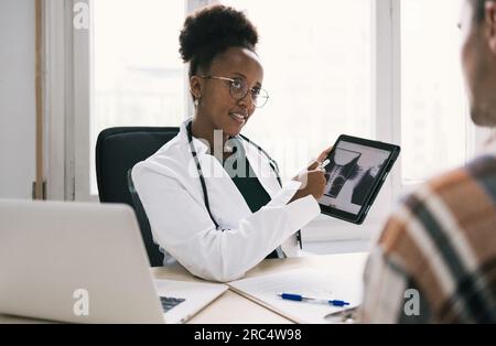 Lächelnde afroamerikanische Ärztin, die Röntgenbilder auf einem Tablet einem nicht erkennbaren männlichen Patienten zeigt und sich ansieht, während sie am Tisch sitzt Stockfoto