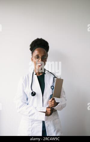 Vorderansicht einer positiven jungen afroamerikanischen Ärztin in Brille, Uniform und Stethoskop, die lächelt und in die Kamera schaut, während sie mit c steht Stockfoto