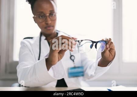 Fokussierte afroamerikanische Ärztin in Brillen, Uniform und Stethoskop, die nach unten schaut, während sie am Tisch sitzt und die Brille mit Bl ansieht Stockfoto