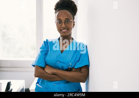 Positive afroamerikanische Ärztin in blauer Uniform und Brille, die lächelt und in die Kamera schaut, während sie steht und sich an die Schulter an der Wand lehnt Stockfoto