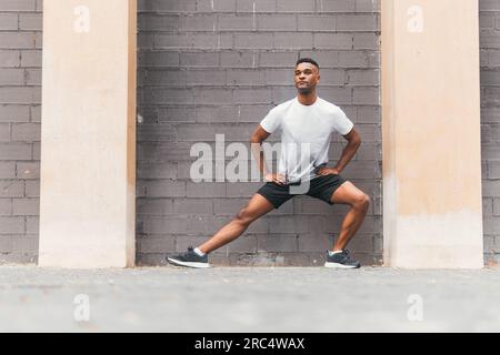 Der ganze Körper besteht aus schwarzem männlichen Sportler in Sportbekleidung und Sneakers, die sich die Beine auf der Straße gegen eine Ziegelwand strecken und wegschauen Stockfoto