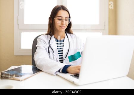 Junge Ärztin in medizinischer Uniform und Brille sitzt am Tisch mit Laptop und gibt Online-Beratung in der Praxis Stockfoto