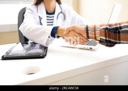 Gekürzte, nicht erkennbare junge Ärztin in Brille und einheitlich schüttelnde Hand eines anonymen Kranken in der Sprechstunde einer modernen Klinik Stockfoto