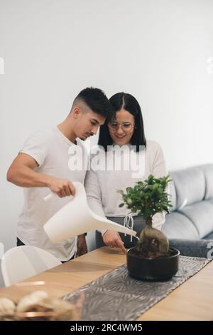 Ein junges Paar in legerer Kleidung, eine Bewässerungspflanze, die auf einem Tisch neben dem Sofa im Wohnzimmer vor verschwommenem Hintergrund platziert wurde Stockfoto