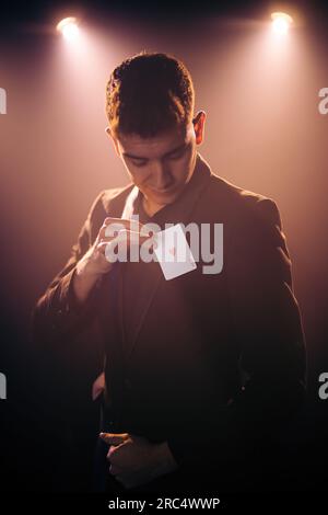 Männlicher Illusionist im Anzug, der einen Zaubertrick zeigt, mit einem Herzchen-Ass, der Karte spielt, während er auf der Bühne gegen die dunkle Wand tritt Stockfoto