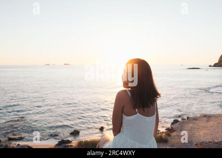 Rückblick auf eine glückliche Frau, die lächelt und über die Schulter in die Kamera schaut, in der Nähe des Wellenmeers im Sommerurlaub auf den Balearen bei Sonnenuntergang Stockfoto