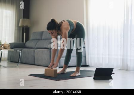 Das ganze Body einer schlanken jungen Frau in Sportbekleidung, die im Online-Unterricht zu Hause die halbe Vorwärtsbeugung mit Yoga-Schwarz in der Nähe des Tablets trainiert Stockfoto