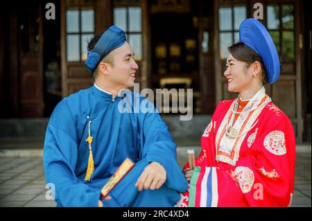 Porträt eines jungen, fröhlichen asiatischen Paares in herrlicher traditioneller Kleidung, das sich ansieht, während es auf der Außenterrasse des traditionellen Hauses sitzt Stockfoto