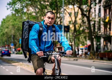 Männlicher Erwachsener mit blauer Jacke, Lieferservice und Rucksack auf Fahrrädern, die Bestellungen in Barcelona ausliefern und wegschauen Stockfoto