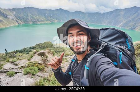 Lächelnder bärtiger männlicher Wanderer mit Hut und Rucksack, der lächelt und in die Kamera schaut, während er Selfie macht und mit einer Handbewegung gegen Quilo auf dem Hügel steht Stockfoto