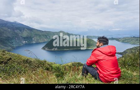 Seitenansicht eines jungen männlichen Touristen, der auf einem Grashügel sitzt und wegblickt, während er die herrliche Landschaft der Cuicocha Lagune Ecuador unter dem bewölkten Himmel in D bewundert Stockfoto