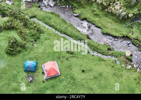 Blick von oben auf farbenfrohe Zelte, die auf einem grasbewachsenen Boden in der Nähe von fließendem Wasser mit grünen Bäumen am Ufer errichtet wurden, am Sommertag in Ecuador Stockfoto