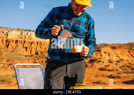 Kulturell positiver reifer Mann in lässiger Kleidung und gelber Mütze steht und gießt heißes Getränk in hügeliger Wüste im Canyon Rojo Teruel Spanien Stockfoto