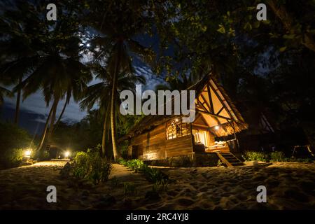 Außenansicht des beleuchteten Hotels mit Holzdach, umgeben von Palmen am Strand bei Nacht in Madagaskar, Afrika Stockfoto