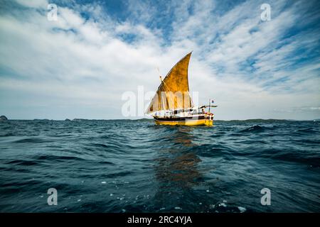 Fischerboot, das auf rauschenden Meerwasser mit offenem gelbem Segel an Deck mit Seilen gefesselt ist, während es an einem sonnigen Tag vor blauem, wolkigen Himmel festgemacht ist Stockfoto