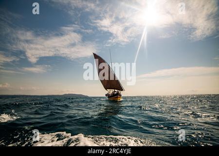 Fischerboot, das auf rauschenden Meerwasser mit offenem gelbem Segel an Deck mit Seilen gefesselt ist, während es an einem sonnigen Tag vor blauem, wolkigen Himmel festgemacht ist Stockfoto