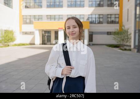 Lächelndes junges Mädchen in Uniform und mit Rucksack, das in die Kamera schaut, während es auf dem Schulgelände neben einem verschwommenen Glasgebäude bei Tageslicht steht Stockfoto