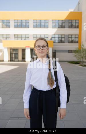 Ein seriöses junges Mädchen in Uniform und mit Rucksack, das in die Kamera schaut, während es auf dem Schulgelände neben einem verschwommenen Glasgebäude bei Tageslicht steht Stockfoto