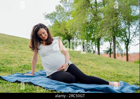 Voller Körper voller Freude, eine Frau in stilvoller Umstandsbekleidung mit großem Bauch zu erwarten, während Sie sich auf einer Decke auf grünem Gras entspannen Stockfoto