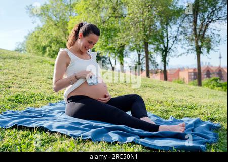 Die ganze Länge einer glücklichen erwartenden Frau mit kleinen Schuhen für das ungeborene Baby auf großem Bauch, während sie auf grünem Gras chillt Stockfoto