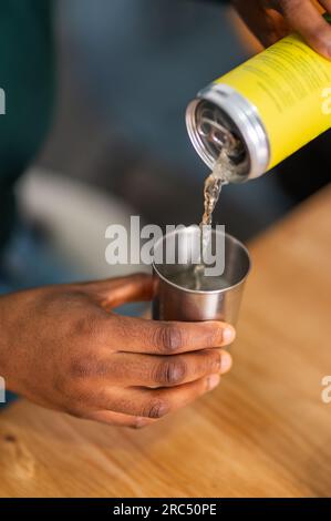 Hoher Winkel des Ernteguts anonyme schwarze Person gießt farbloses Natronlauge aus einer Dosenflasche in einen Edelstahlbecher, während sie auf einem Holztisch sitzt Stockfoto