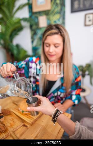Eine junge lächelnde Frau in legerer Kleidung sitzt am Holztisch und gießt Wasser aus der Kanne aus Edelstahl mit geernteter Hand in ein helles Restaurant Stockfoto