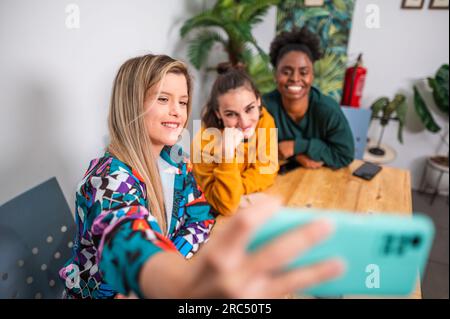 Eine Gruppe von fröhlichen, multirassischen Freunden in lässiger Kleidung, die in der Cafeteria am Tisch sitzen und Selfie auf dem Smartphone machen Stockfoto