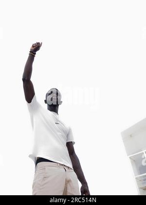 Von unten, von einem seriösen, selbstbewussten afroamerikanischen Kerl in lässiger Kleidung, der auf der Straße der Stadt steht, mit erhobenem Arm und Finger nach oben Stockfoto