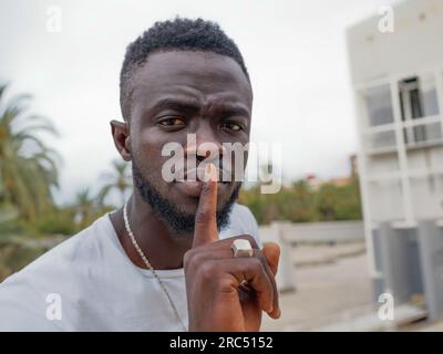 Selbstbewusstes junges bärtiges afroamerikanisches männliches Model in lässiger Kleidung, das Stille zeigt und im Stadtpark vor die Kamera schaut Stockfoto