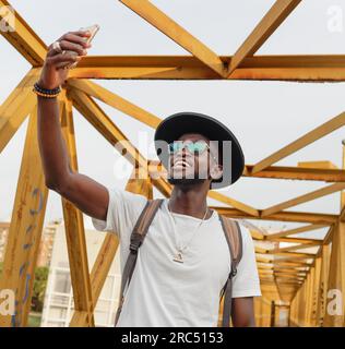Cooles, gutaussehendes Männchen mit Hut und Rucksack, der auf der Brücke steht und Selfie auf dem Handy macht Stockfoto