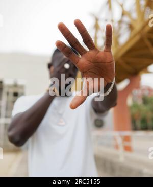 Nicht wiedererkennbarer junger afroamerikanischer Mann in weißem T-Shirt, der das Gesicht bedeckt und die Hand in Richtung Kamera streckt, während er auf der Straße der Stadt steht Stockfoto